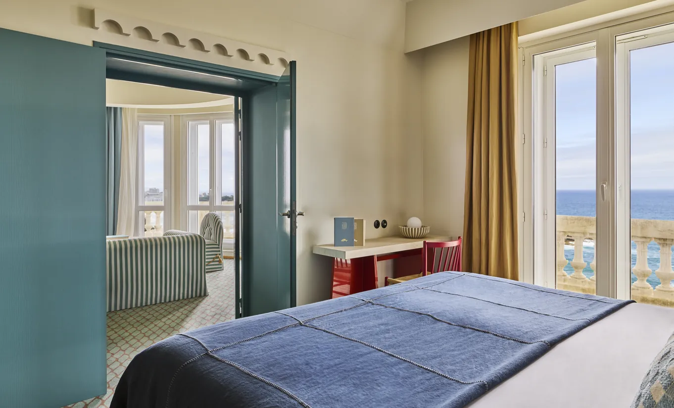 Experimental Group hotel bedroom with blue headboard, white bedding, teal bathroom door ajar, and large window overlooking the sea