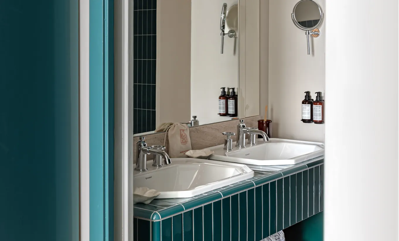 Experimental Group hotel bathroom with teal green vanity unit, marble double sink, brushed brass fixtures, and white tiled walls