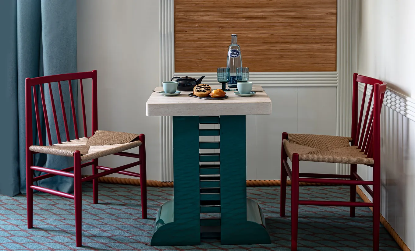 Experimental Group hotel room corner with teal storage unit, two wooden woven chairs, and natural light from window with blinds