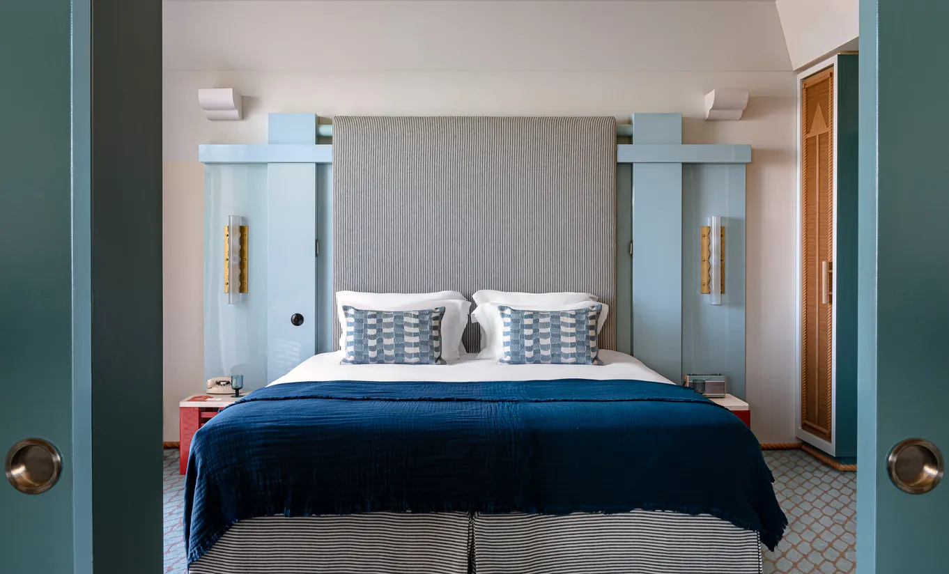 Experimental Group hotel bedroom with grey upholstered headboard, navy blue bedding, teal wardrobe doors, and soft ambient lighting