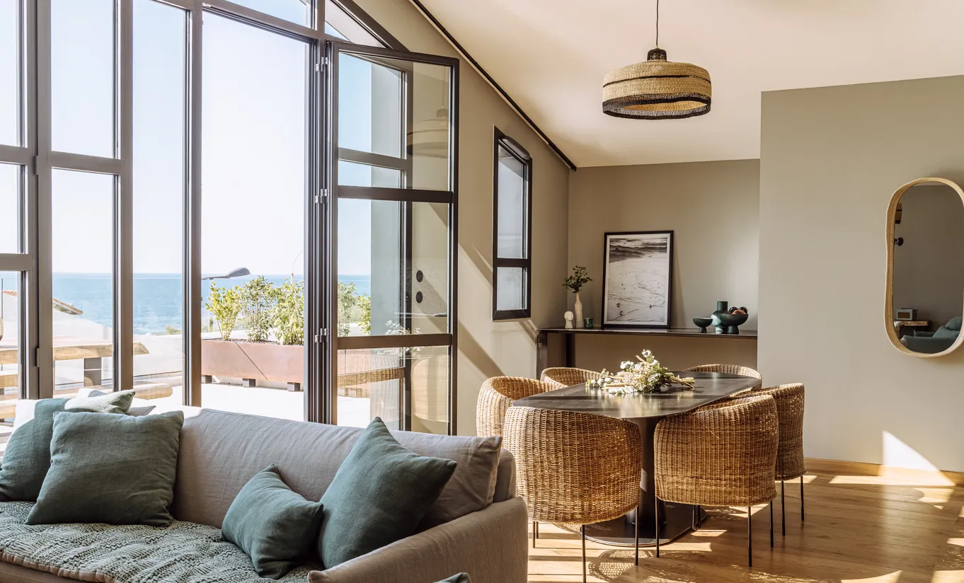Experimental Group hotel sunlit open-plan living room with grey sofa, dining area and large industrial steel-framed windows overlooking rooftops