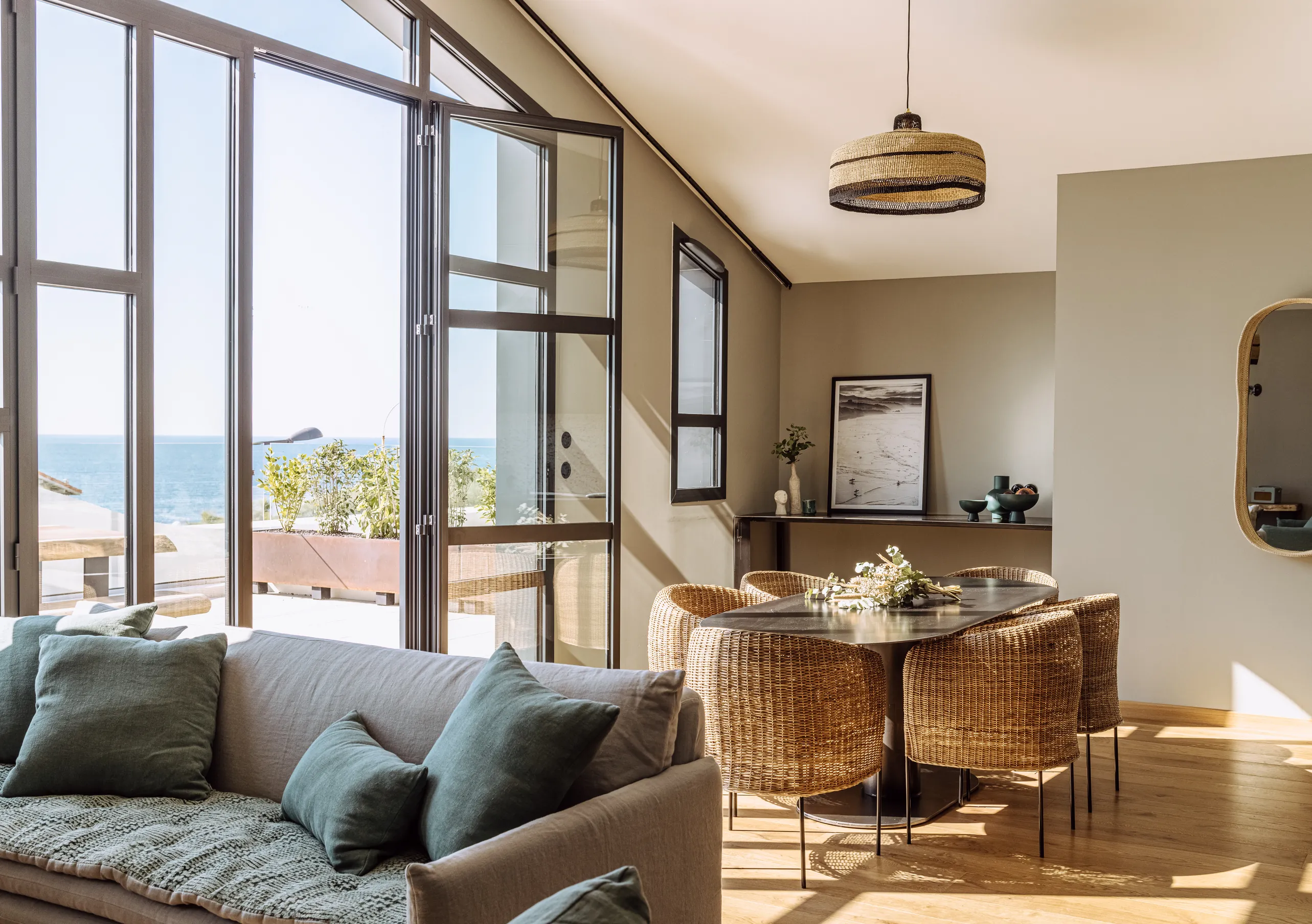 Experimental Group hotel sunlit open-plan living room with grey sofa, dining area and large industrial steel-framed windows overlooking rooftops.