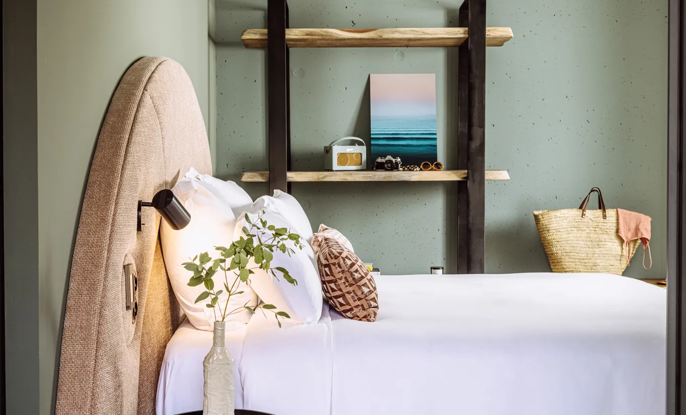Experimental Group hotel bedroom with rounded natural wood headboard, white bedding, potted plant, and open shelving with decorative items