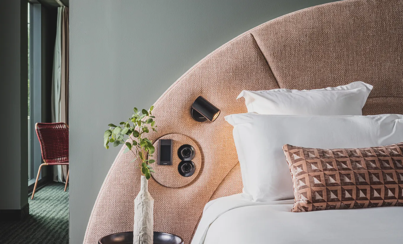 Experimental Group hotel bed detail with curved blush pink headboard, embroidered cushions, integrated wooden bedside shelf, and warm lighting