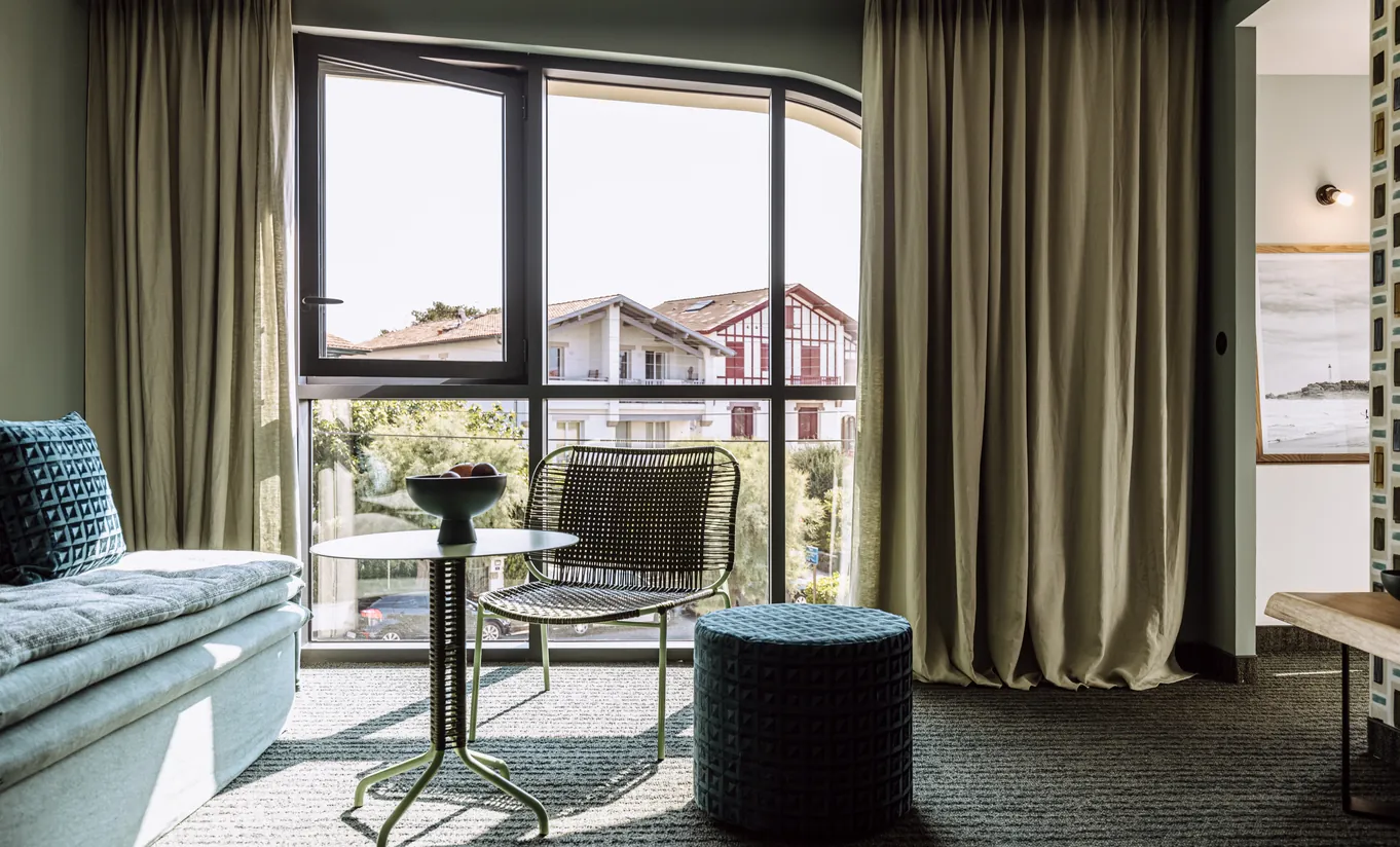 Le Garage Biarritz Experimental hotel room with beige linen curtains, work desk, metal chair, and large window with urban city view