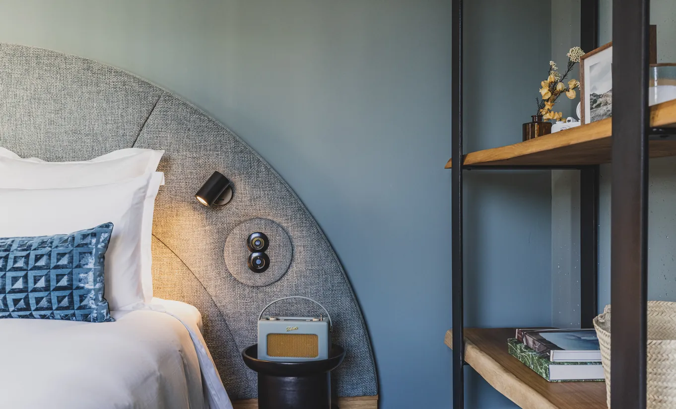 Le Garage Biarritz Experimental hotel bedroom detail with curved headboard, blue geometric cushion, wall-mounted reading light, and wooden bedside shelf