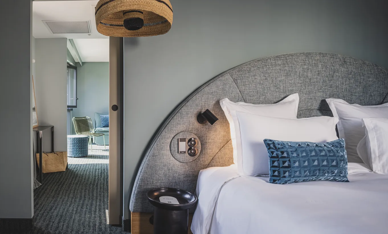 Le Garage Biarritz Experimental hotel bedroom with curved grey headboard, blue patterned cushions, floor lamp, and open bathroom en suite