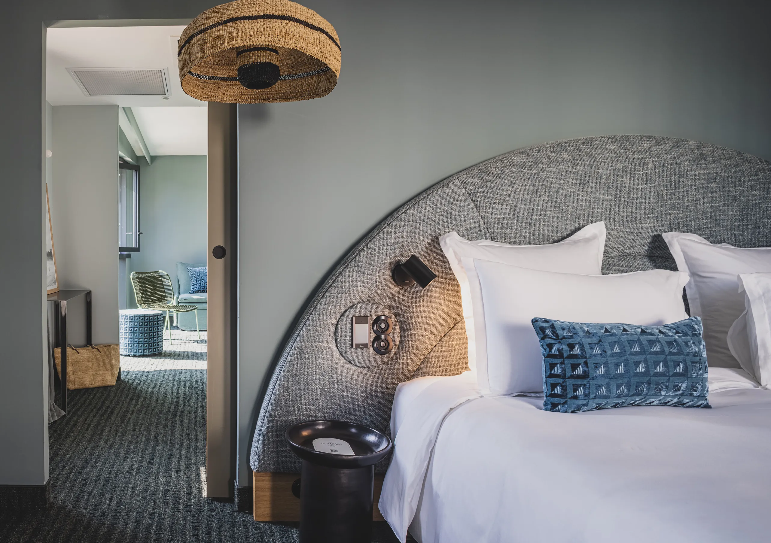 Le Garage Biarritz Experimental hotel bedroom with curved grey headboard, blue patterned cushions, floor lamp, and open bathroom en suite.