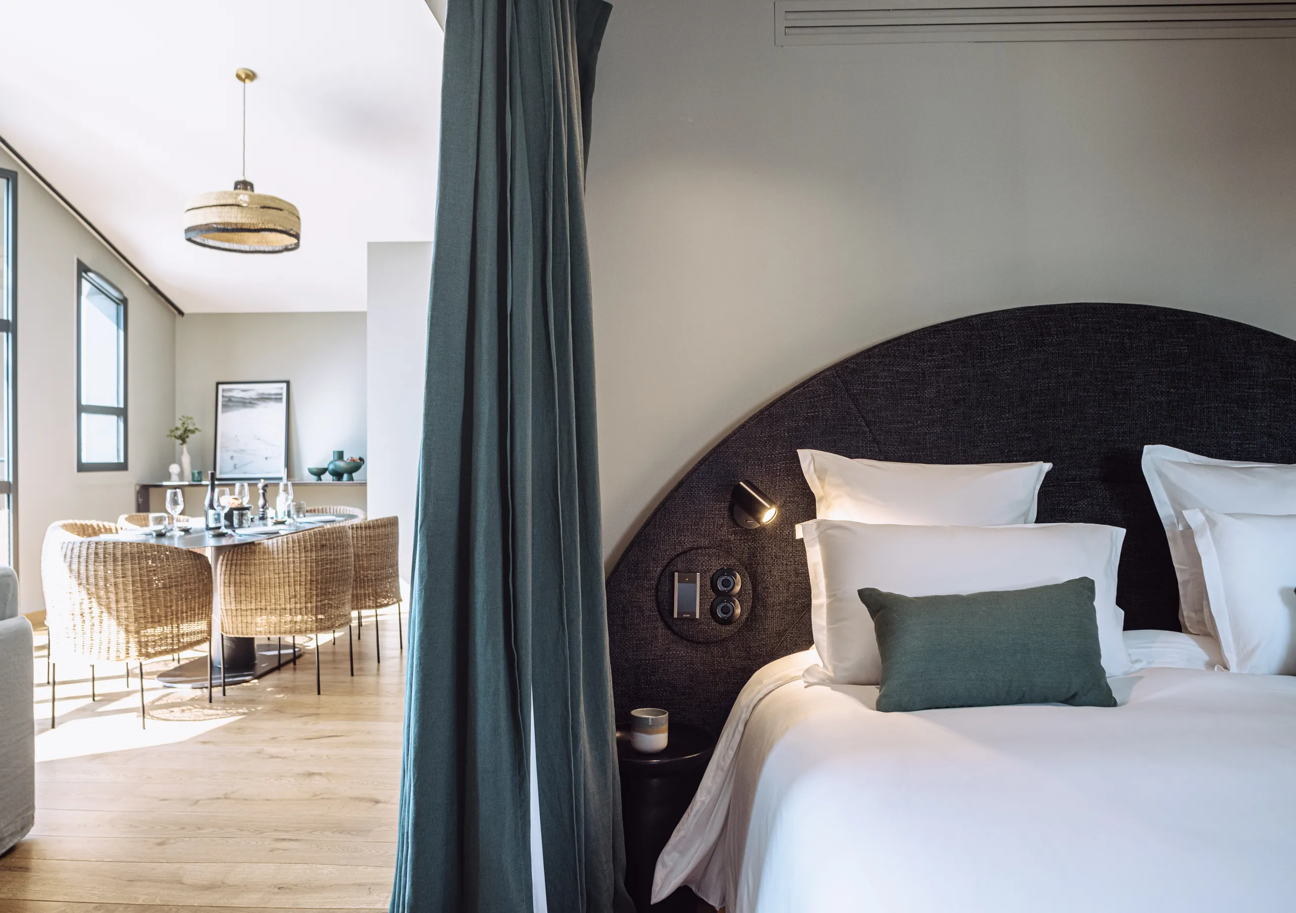 Experimental Group hotel cozy bedroom with dark teal headboard, white bedding and open view to a bright living area.