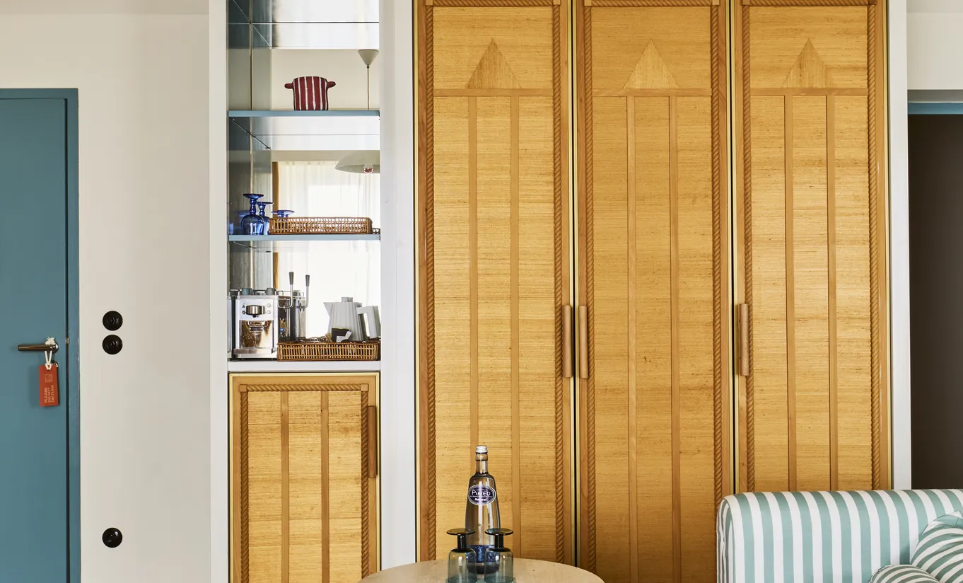 Experimental Group hotel room with tall warm wood built-in wardrobe, open shelving with books, and blue accent door detail