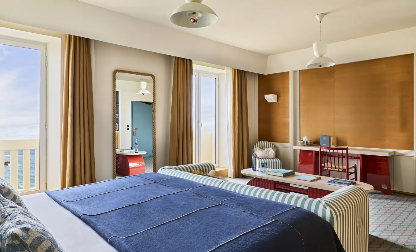 Experimental Group hotel suite with king bed, warm wood paneling, orange curtains, and open view into a bright living area