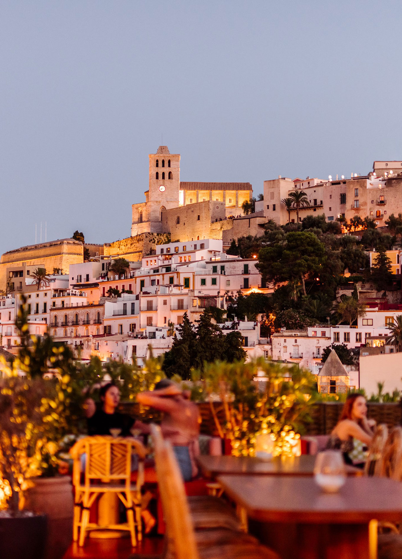 An evening view of a restaurant terrace with tables and guests, overlooking a historic illuminated town on a hillside.