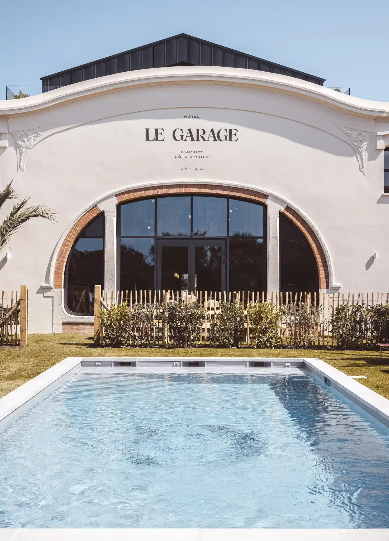Façade du bâtiment « Le Garage » avec une grande fenêtre cintrée et une piscine au premier plan.