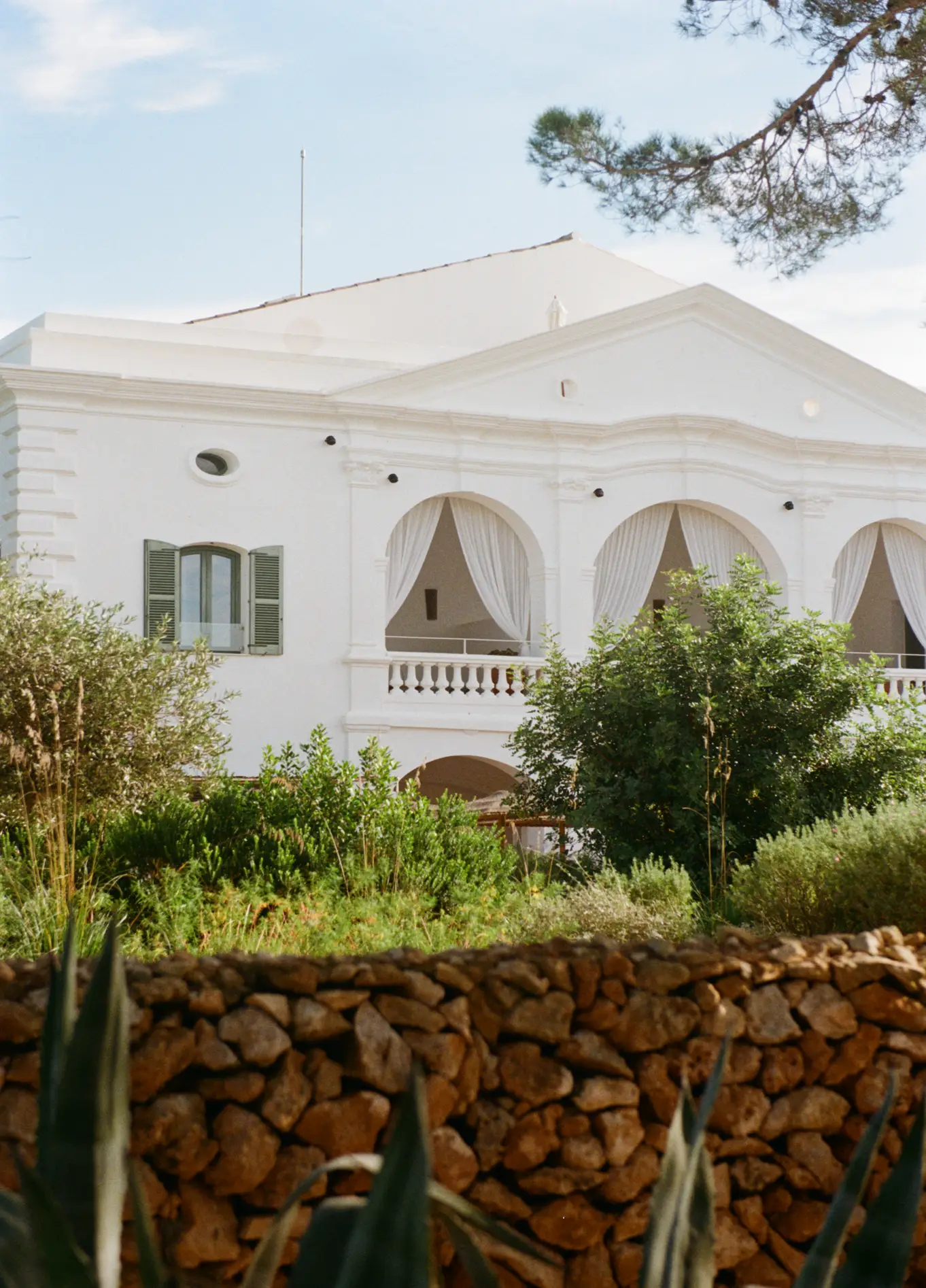 Immaculate white facade of the Menorca Experimental hotel, surrounded by lush vegetation and a dry stone wall in the foreground.