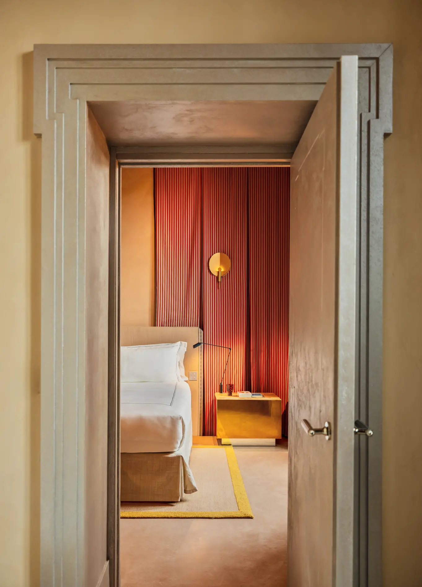 View through an ajar door leading into an elegant bedroom with warm tones, featuring a bed and a gold bedside table visible in the background.