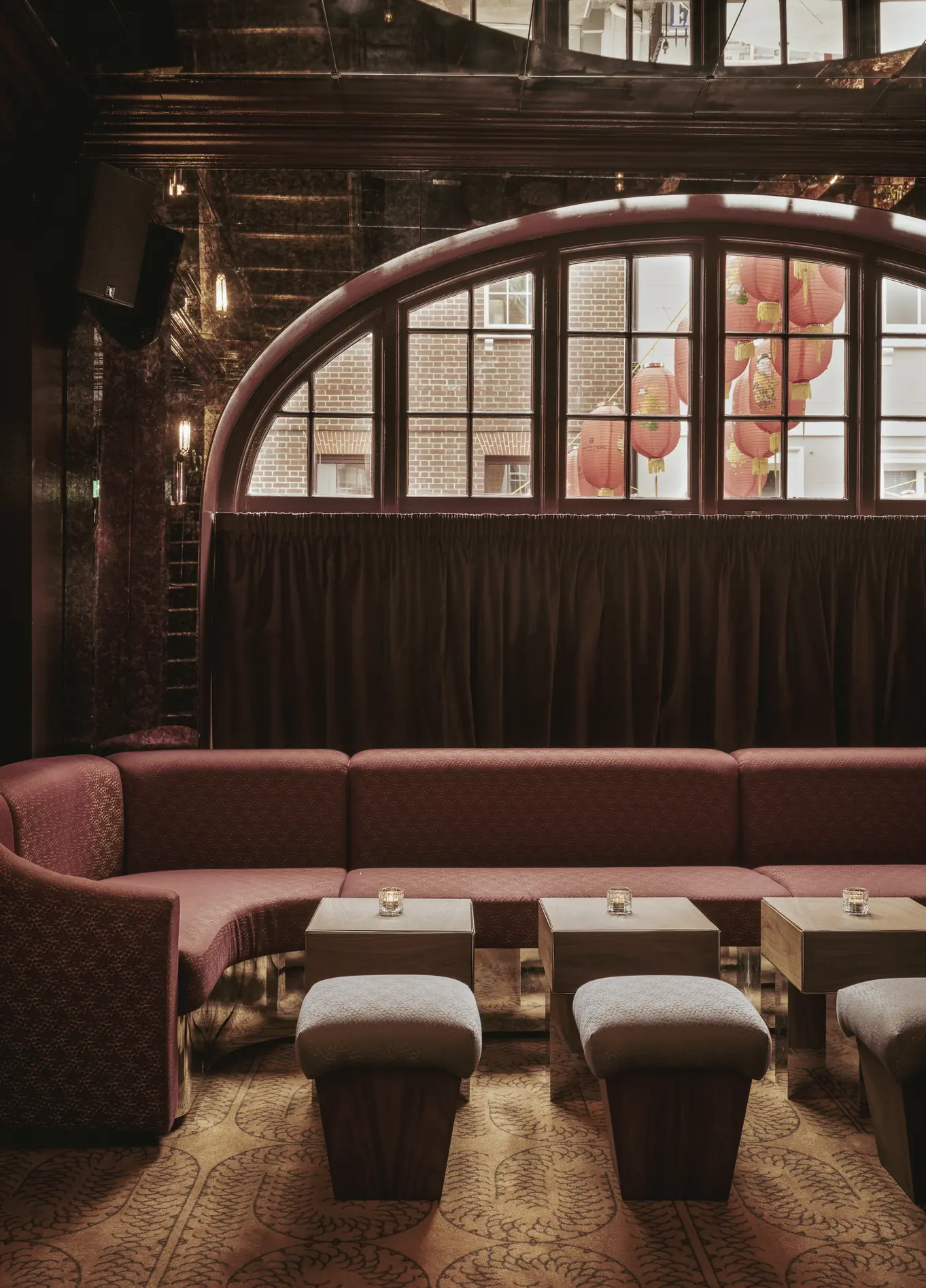 A cozy lounge area with large red sofas and coffee tables in front of a large arched window at the Experimental Cocktail Club.