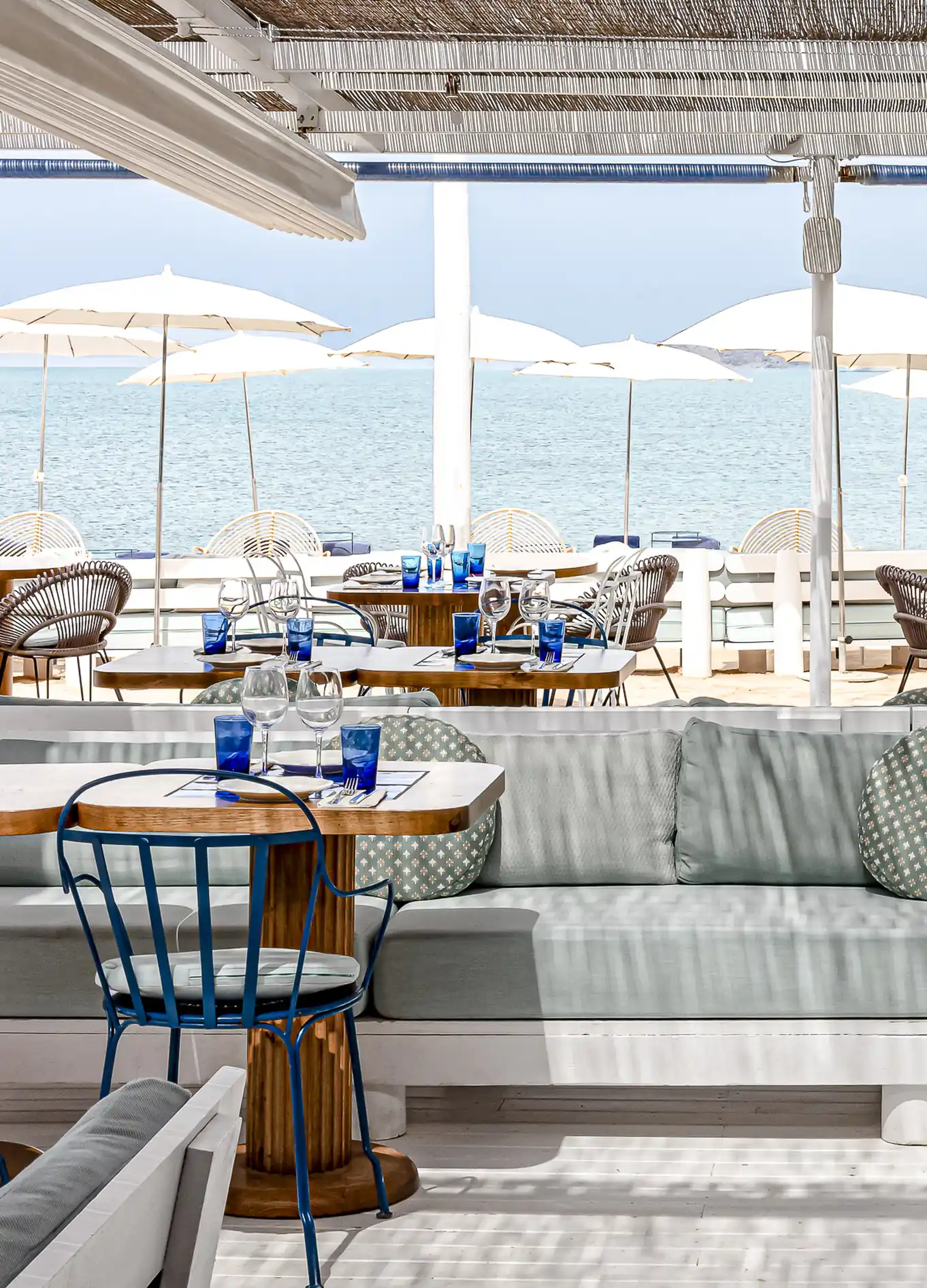 A restaurant terrace with comfortable sofas and tables, offering a view of the sea with many white umbrellas.