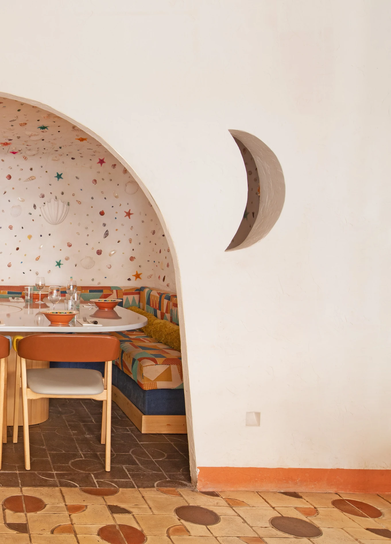 A dining area viewed through an archway, featuring a moon-shaped wall decoration, tables with chairs, and a tiled floor.