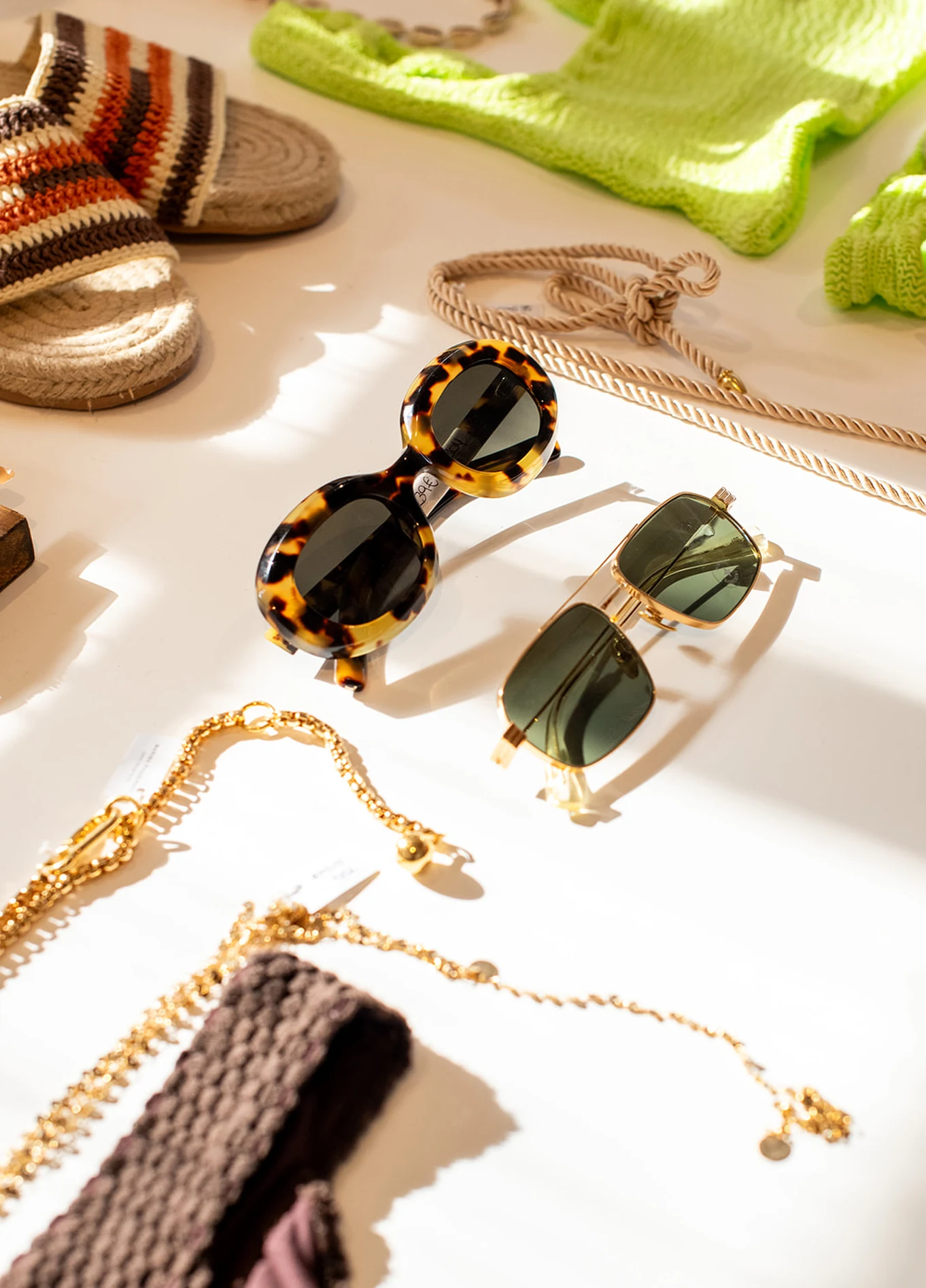 A selection of fashion accessories, including sunglasses, gold jewelry, and shoes, arranged on a light-colored surface.
