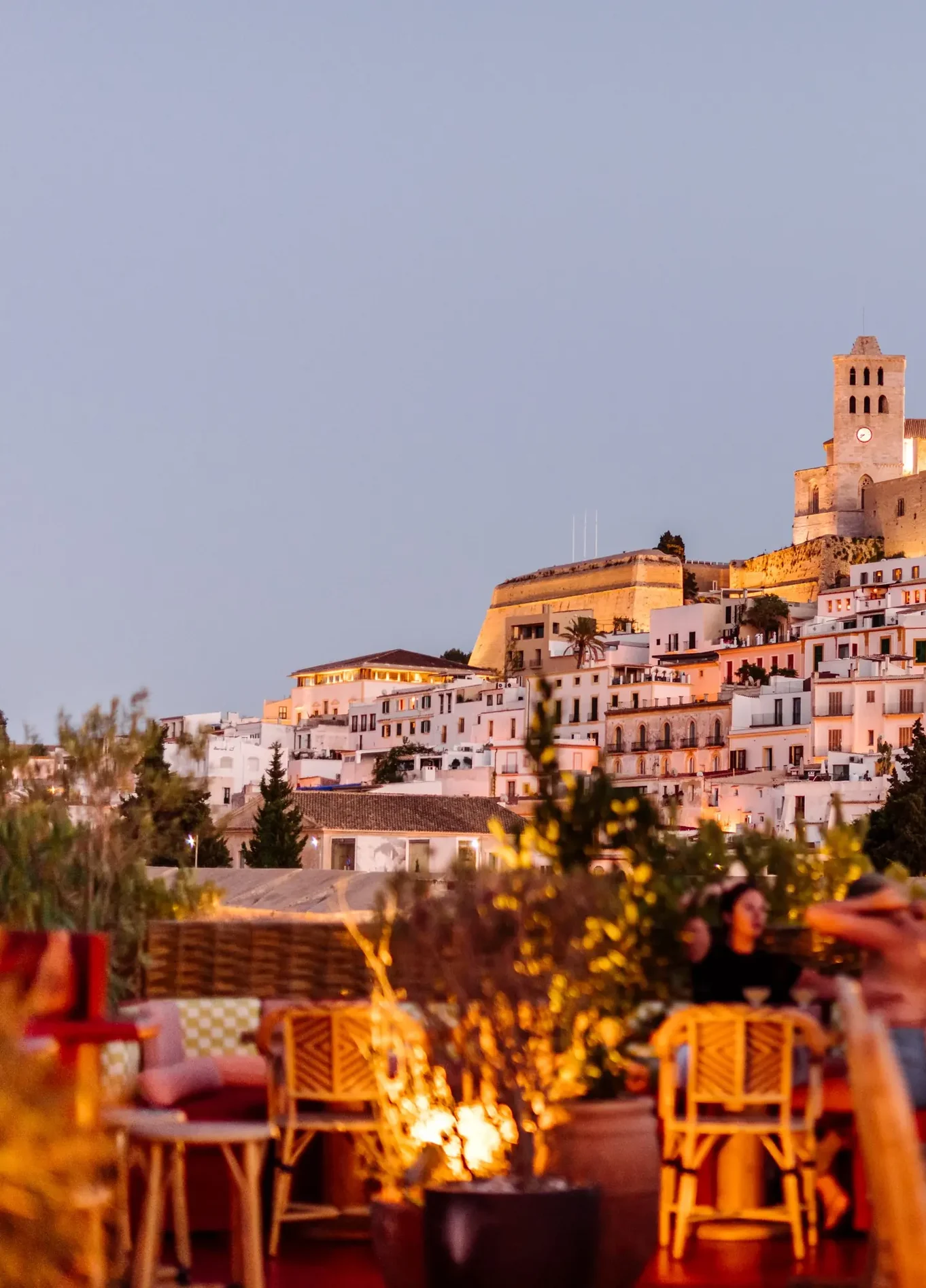 An evening view of a restaurant terrace with tables and guests, overlooking a historic illuminated town on a hillside.